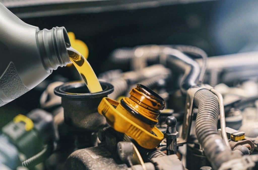 A bottle of oil is being poured into a yellow funnel, which is connected to a car engine. The scene takes place in a garage, with the engine visible in the background. The oil is being poured into the engine to ensure proper lubrication and maintenance.